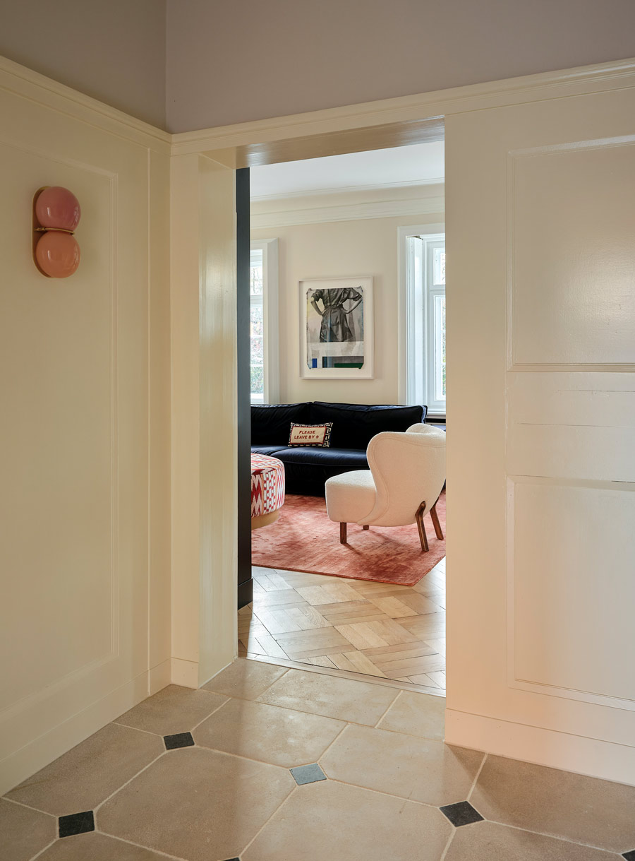  Zu sehen ist hier ein Blick von der Eingangshalle in das Wohnzimmer. Die Eingangshalle hat einen hellen Natursteinboden mit schwarzen Einsätzen, im Wohnzimmer liegt Parkett. Dort ist der Petra Lounge Chair mit Schafsfell zu sehen, ein großes dunkelblaues Samtsofa, ein warmer terrakottafarbenen Viskoseteppich und ein großer, runder Coffee Table aus Stoff mit einem Ikat Muster in Warmen Rot-Tönen.